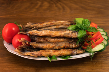 Small fish (capelin), fried until golden  brown, stacked slide on a plate with slices of cucumbers, sweet pepper and whole tomatoes.
