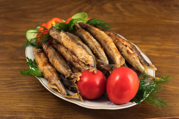 Fried capelin with crispy crust, laid on a porcelain plate with a garnish of fresh vegetables and herbs.
