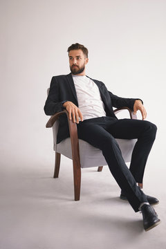 Vertical shot of serious fashionable elegant young bearded male wearing stylish shoes, trousers and trendy jacket over white t-shirt sitting in armchair, having confident bossy facial expression