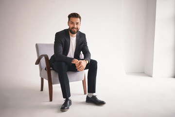 People, lifestyle, business, style, fashion and men's wear concept. Positive successful young CEO sitting in armchair, smiling at camera, dressed in elegant shoes, trousers, jacket and white t-shirt - Powered by Adobe