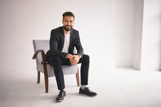 People, lifestyle, business, style, fashion and men's wear concept. Positive successful young CEO sitting in armchair, smiling at camera, dressed in elegant shoes, trousers, jacket and white t-shirt