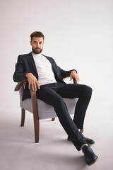 Isolated vertical portrait of successful handsome stylish young European male boss with fuzzy trimmed beard wearing trendy men's wear relaxing in armchair and staring at camera with serious look