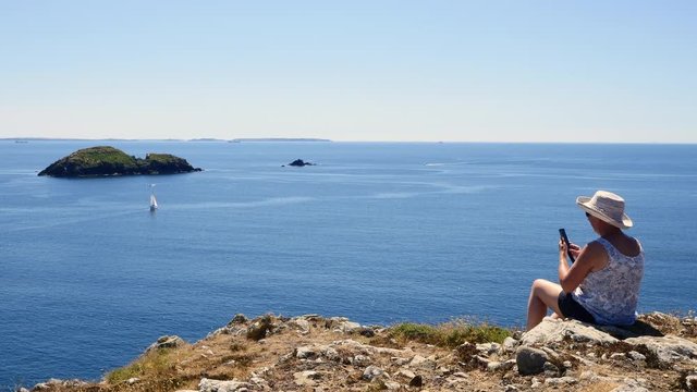Lady on a cliff top, with a phone camera caputuring the view across the bay.