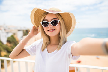 Fototapeta premium Beautiful woman in sunglasses and summer hat taking selfie on beach