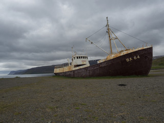 Fototapeta premium Iceland, west fjords, Patreksfjordur, June 24, 2018: Old abandoned rusty fishing boat ship wreck standing on the sand beach coast in west fjords od Iceland, dramatic gray sky and cliffs background