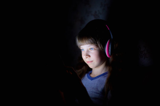 the child in wireless headphones listens to music at night under a blanket and looks at the tablet