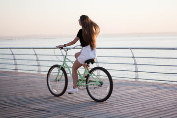 Fototapeta premium Beautiful longhaired young woman with a bicycle having fun, cycling, posing on the camera near the sea on the sunrise