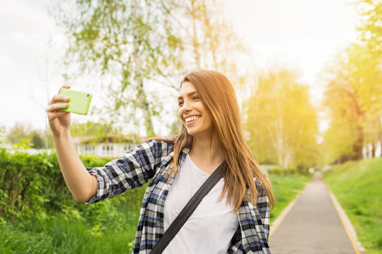 Young woman taking a selfie or filming a vlog video on smartphone outdoors in park. Beautiful blonde teenage girl filming or photographing herself outside on sunny day.