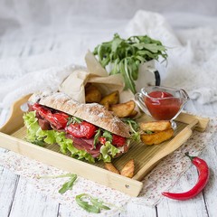 sandwich with ham, cream cheese, lettuce leaves, arugula, red pepper, baked potatoes, ketchup on a white wooden background with a floral fabric