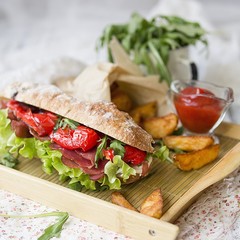 sandwich with ham, cream cheese, lettuce leaves, arugula, red pepper, baked potatoes, ketchup on a white wooden background with a floral fabric