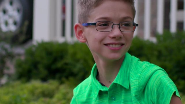 A cute boy hanging out in the front yard looks up at someone coming down the street off camera and smiles and waves hi in Slow Motion.