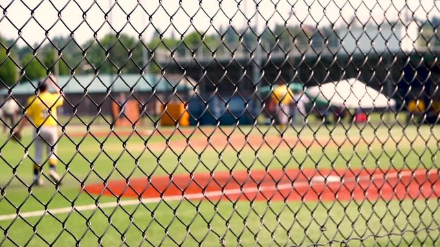 A pitcher in a youth baseball game throws a pitch, resulting in an out at first base. Game action is viewed through a chain link fence, with selective focus on the fence with the players out of focus.