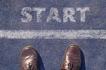 man feet in brown leather shoes standing before shabby sign start written on asphalt, top view