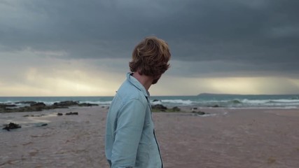 A man standing on a beach with his back to the camera looking out to see with light rays shining through storm clouds in slow motion. - Powered by Adobe