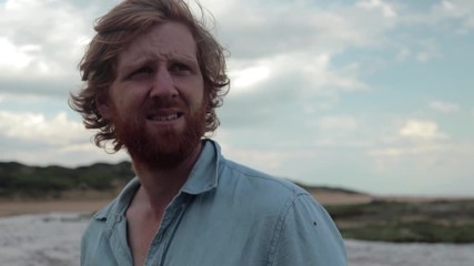 A slow motion close shot orbiting around a long haired ginger man standing on the beach looking out to sea. - Powered by Adobe