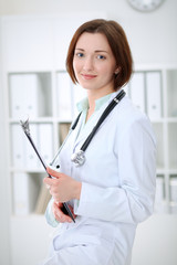 Young brunette female doctor standing with clipboard and smiling in hospital