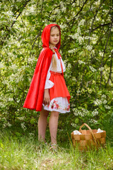 Smiling blond girl posing in a dress of little red riding hood near green trees