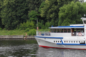 Fototapeta na wymiar Bow of white tourist ship slowly floating along the concrete coast of the navigable Moscow canal in the summer against the green trees on the shore, side view – travel, tourism, water recreation
