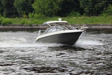 Travel, luxury water recreation on yacht - a white 19 ft motor boat with hardtop fastly floating along the concrete shore of the navigable canal of Moscow in summer amid the trees on shore, side view