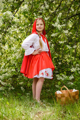 Smiling blond girl posing in a dress of little red riding hood near green trees