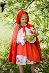 Smiling blond girl posing in a dress of little red riding hood near green trees