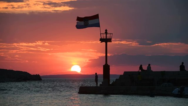 Stone pier with lighthouse and flying Croatian flag in sunset time lapse, people walking and sitting on stone wall, silhouettes of people only, individuals not recognizable