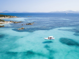View from above, aerial picture of a boat with some relaxed tourists on board floating on a transparent and turquoise Mediterranean sea. Emerald Coast (Costa Smeralda) in Sardinia, Italy.