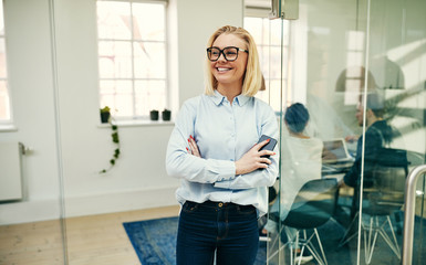 Smiling businesswoman leaning with her cellphone against an offi