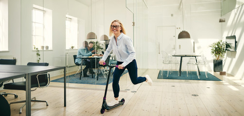 Laughing young businesswoman riding a scooter in a large office