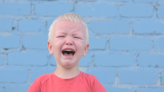 Little blond boy in red T-shirt is crying with tears against gray brick wall.
