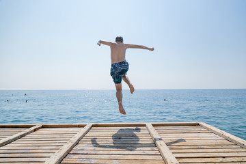 Fototapeta na wymiar Teenage boy, jumping into the sea from wooden jetty on a beach. Happy summer vacation concept.