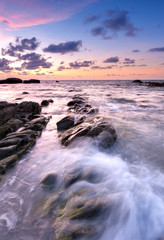 Obraz premium view of beautiful sunset at the beach with natural coastal rocks covered by green moss. soft focus due to slow shutter effect.