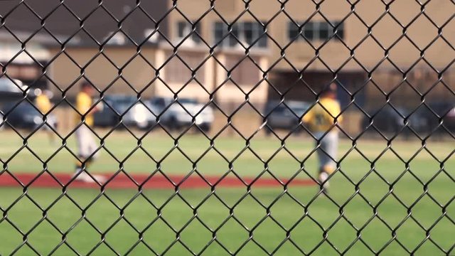 A shortstop fields a ground ball and throws to first base. A runner runs to thirds base. Game is viewed through a chain link fence, with selective focus on the fence with the players out of focus.