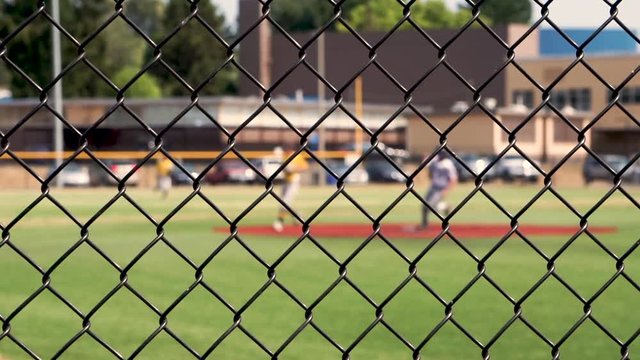 Two runners round second base past the shortstop and second baseman after the batter hits a triple. Viewed through a chain link fence, with selective focus on the fence with the players out of focus.