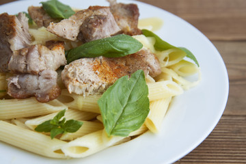 Cooked penne pasta isolated in white round plate, with basil leaves and pork meat, over wooden table, decorated with basil leaves. Top view.