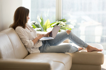 Calm female working at laptop sitting on cozy sofa, happy girl browsing internet or shopping online...