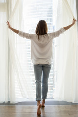 Fototapeta na wymiar Back view of casual woman opening curtains enjoying beautiful sunny morning, happy female looking out from apartment window, admiring skyscrapers outline, waking up starting day positively