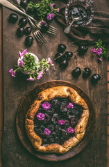 Homemade open cherry pie or galette on wooden rustic kitchen table background with jam, flowers and cutlery, top view. Summer berries still life concept