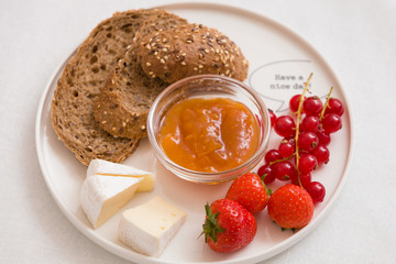 Top view on white plate with slices of organic wholegrain bread, Camembert cheese, apricot jam, red current and strawberries. Healthy snack or breakfast at home.