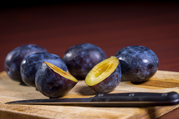 Fresh plums and one sliced plum with knife on a wooden board on dark red table