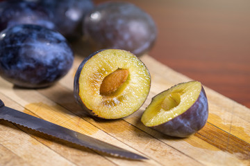 Fresh plums and one sliced plum with knife on a wooden board on dark red table