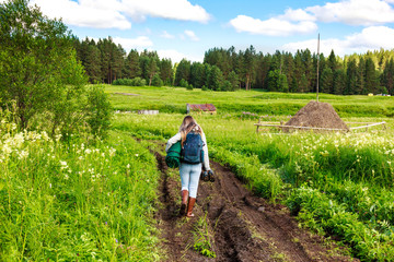  A girl is walking along a rural road in rubber boots with a backpack and sleeping bag