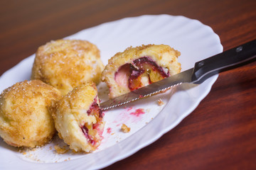 Fresh plum dumplings. One sliced dumpling and two whole with knife and fork on white plate on dark red table