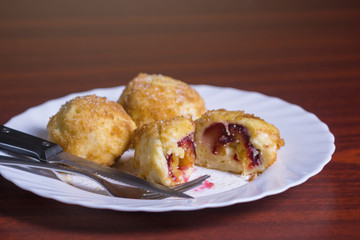 Fresh plum dumplings. One sliced dumpling and two whole with knife and fork on white plate on dark red table