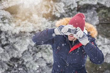 Fashionable man taking pictures with retro camera on snowy day. Winter vacation travel concept.