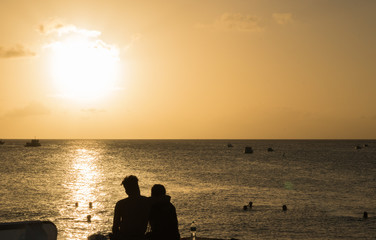 Fototapeta na wymiar A young couple in love sits in an embrace and looks at the sunset over the horizon of the sea, swimming people and fishing boats in the distance. Silhouettes. Concept: Holiday together.