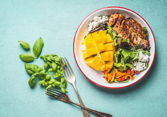 Tasty summer salad with roasted chicken breast and mango in bowl with cutlery on light blue...