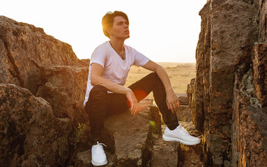 guy sits on a rock in a t-shirt, sneakers and jeans in the summer, sunset