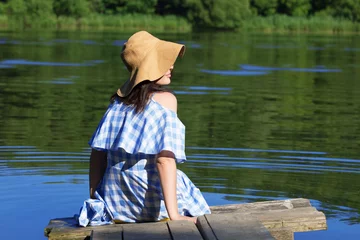 Foto op Canvas Happy young woman sitting on the bridge near the river. A blue dress and a straw hat. Back view. © AnastazjaSoroka