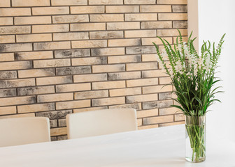 Vase with beautiful flowers on white table against brick wall. Stylish interior